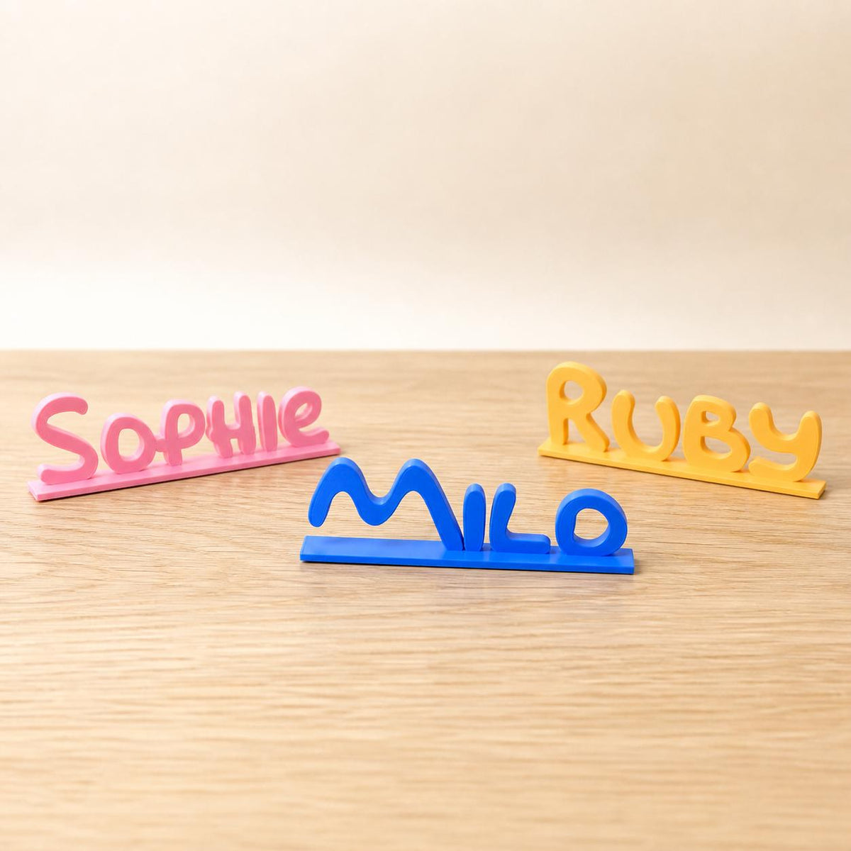 Sophie, Milo and Ruby cartoon style personalised PLA nameplates arranged in a warm neutral studio setting.