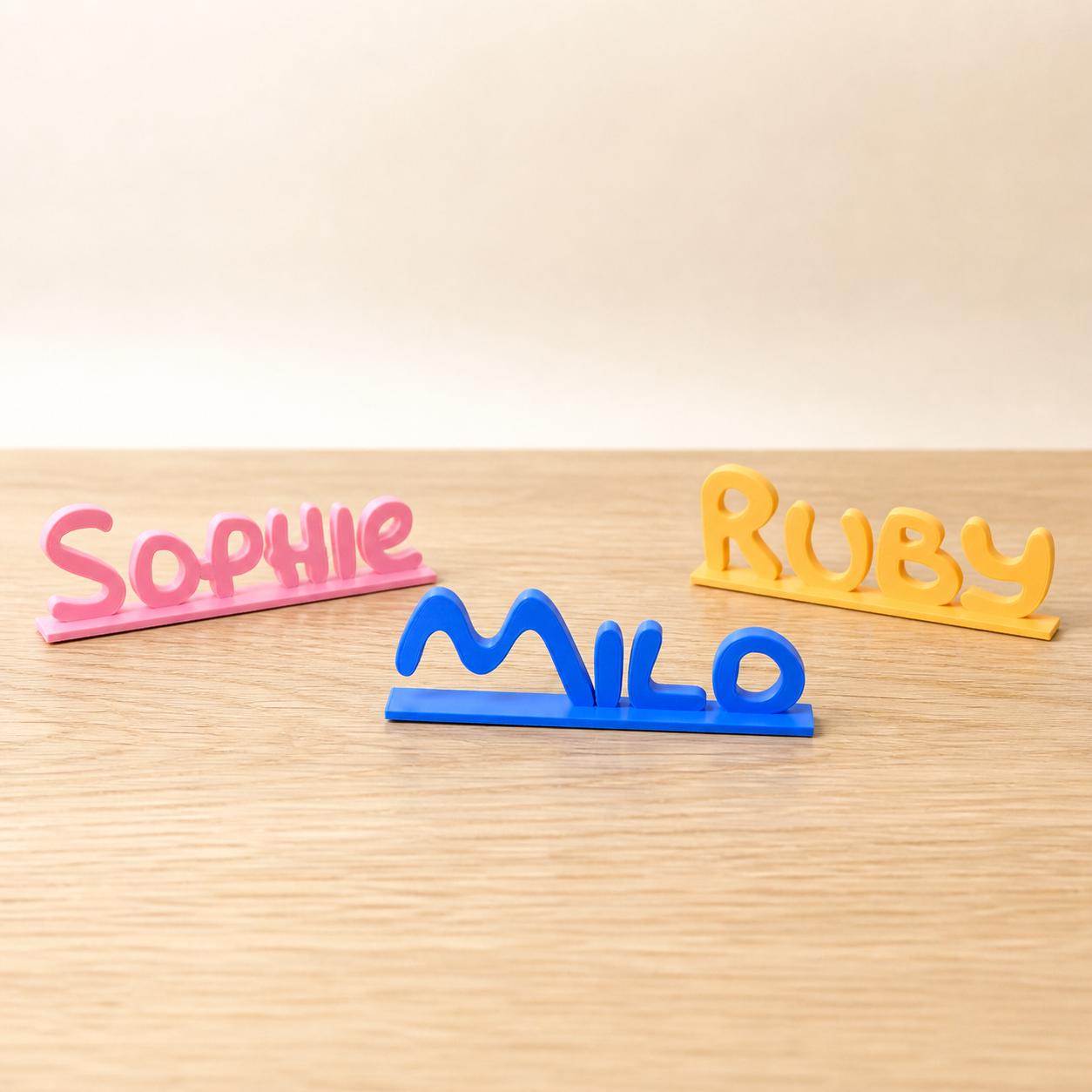 Sophie, Milo and Ruby cartoon style personalised PLA nameplates arranged in a warm neutral studio setting.