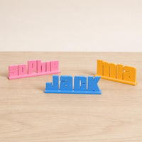 Sophie, Jack and Mia bold arcade personalised PLA nameplates arranged in a warm neutral studio setting.